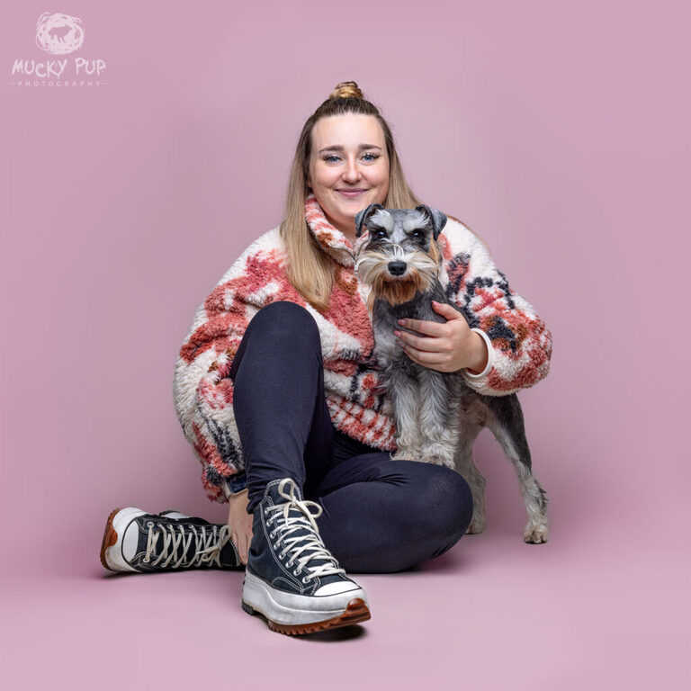 A miniature schnauzer dog and her female owner photographed in a studio setting on a pink background. 